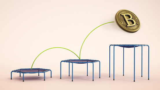 A conceptual visualization presenting a large, gold and black Bitcoin coin suspended in the air, directly above a blue trampoline with a purple surface, set against a plain pink backdrop, hinting at the volatility of cryptocurrency markets.