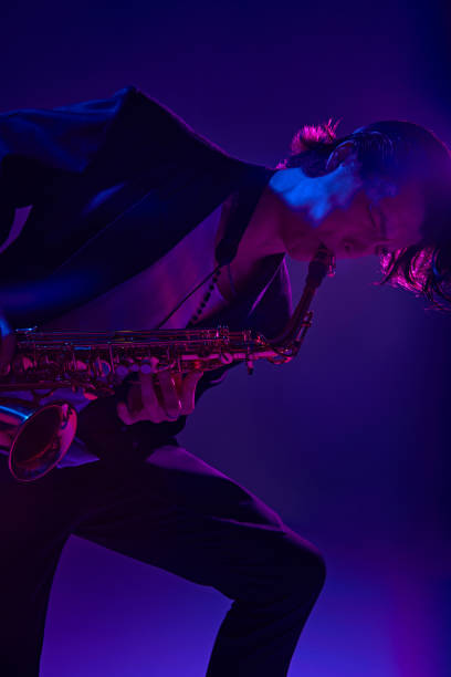 istock Young virtuoso in classical black suit playing saxophone in vibrant pink neon light against dark studio background. 2149263678