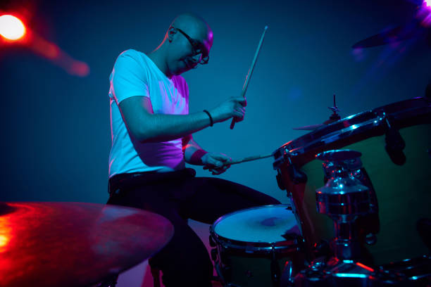 istock Focused bald drummer in white shirt playing in pink-purple stage lighting against gradient studio background. 2149263714