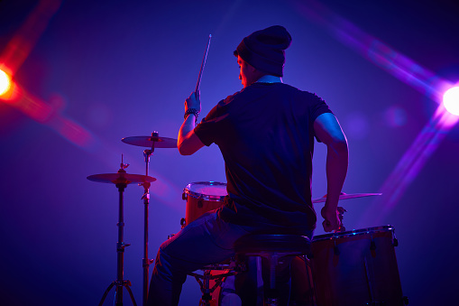 Rear view portrait of young artistic man playing drums in pink-purple stage lighting against gradient studio background. Concept of music and art, hobby, concerts and festivals, modern culture. Ad