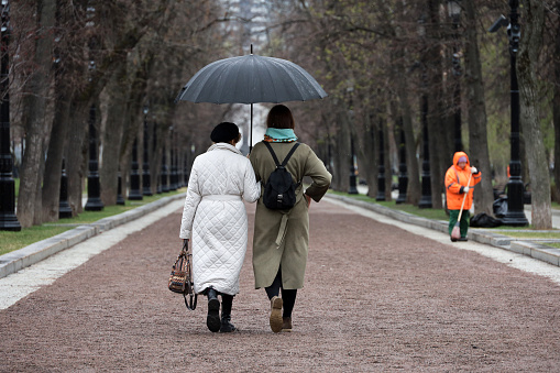Moscow, Russia - April 18, 2024: Two women with one umbrella walking on city street. Rainy weather in spring park