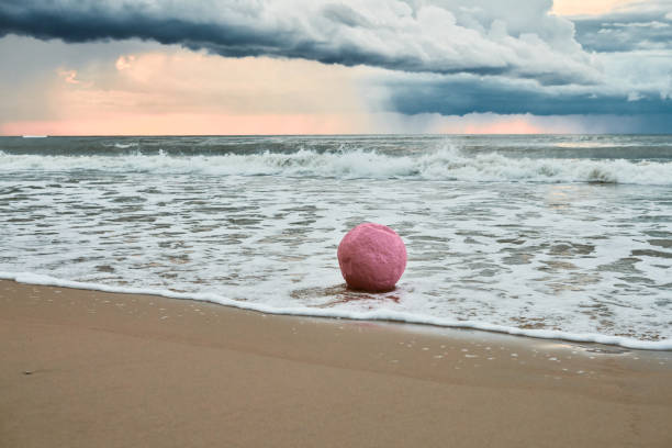 Large pink sphere lapped by waves against seashore against horizon and storm clouds, pink ball symbol of serenity contrasts against brewing turbulence in sky and relentless energy of sea