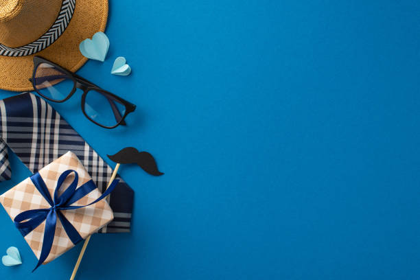 istock Show Dad your love and appreciation with tasteful top view setup featuring straw hat, stylish necktie, adorned gift box, glasses, mustache, and paper hearts on blue backdrop, perfect for greeting text 2149267182