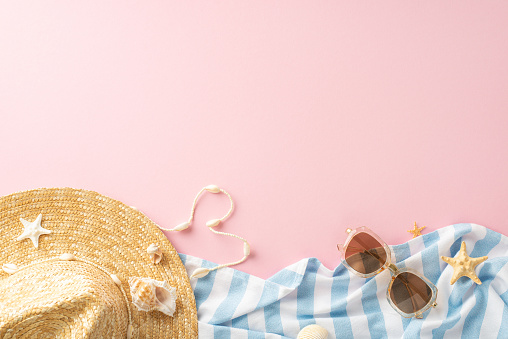 Top view of summer vacation accessories with a straw hat, sunglasses, and a striped towel adorned with seashells and starfish on a vibrant pink background