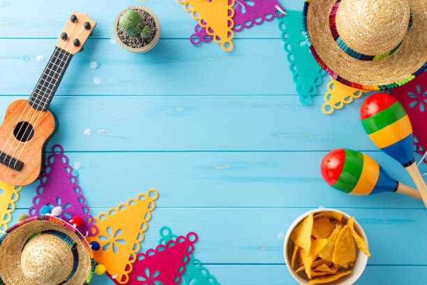 istock Mexican-themed arrangement for Cinco de Mayo. Overhead shot displaying cultural items: sombreros, vihuela, maracas, cactus, flag garlands, and nachos on blue wooden surface. Ideal for event promotions 2149267268