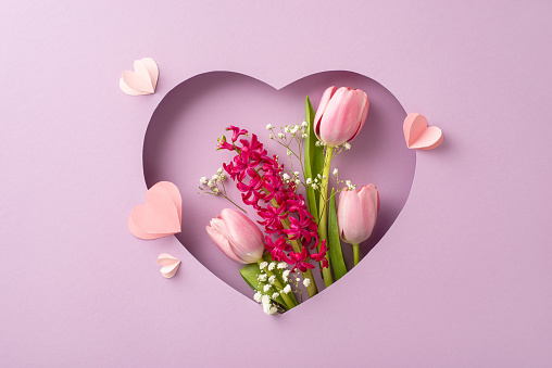 Honor Mom: Top view of fresh tulips, hyacinth, gypsophila, paper hearts elegantly displayed within heart-shaped cutout frame against pastel lilac background. Great for personalized messages or adverts
