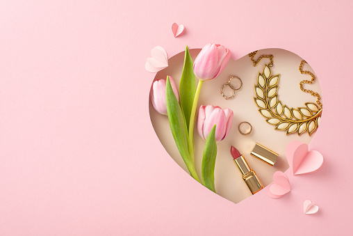 Sophisticated Mother's Day presentation: Top view of tulips, lipstick, gold jewelry, necklace, rings, and paper hearts in heart-shaped frame on soft pink surface, with text space for greetings or ads