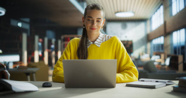 istock Focused Young Teacher Preparing for Class in a Library Reading Hall. Successful Female Using Laptop Computer to Work on a University Research Project, Writing Down Ideas in Notebook 2149269810