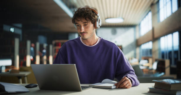 istock Handsome Student Writing an Essay on Laptop Computer, Thinking About the Topic, Brainstorming, Finding Solutions. Young Stylish Man Studying in a Public Library, Listening to Lecture in Headphones 2149269876