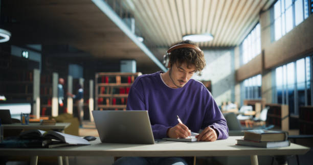 handsome student writing an essay on laptop computer, thinking about the topic, brainstorming, finding solutions. young stylish man studying in a public library, listening to lecture in headphones - studera bildbanksfoton och bilder