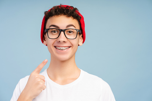 Smiling teenage boy with braces wearing stylish red hat and eyeglasses, pointing finger at his teeth, looking at camera, standing isolated on blue background. Dental care, treatment, health care