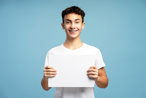 Handsome smiling boy, teenager with braces holding white blank screen, copy space, looking at camera standing isolated on blue background. Advertisement concept
