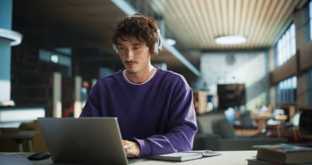 istock Handsome Student Writing an Essay on Laptop Computer, Thinking About the Topic, Brainstorming, Finding Solutions. Young Stylish Man Studying in a Public Library, Listening to Lecture in Headphones 2149269958