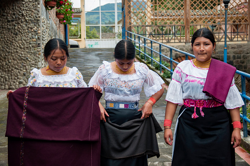 Three women dressed in cultural attire display their weaving skills on a village road in Ecuador.