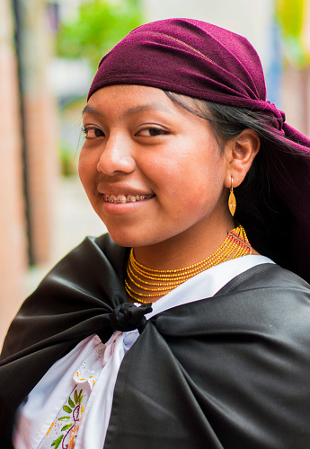 An Ecuadorian woman with a striking purple scarf on her head and a wide black cape stands with an air of mystery and elegance looking at the camera