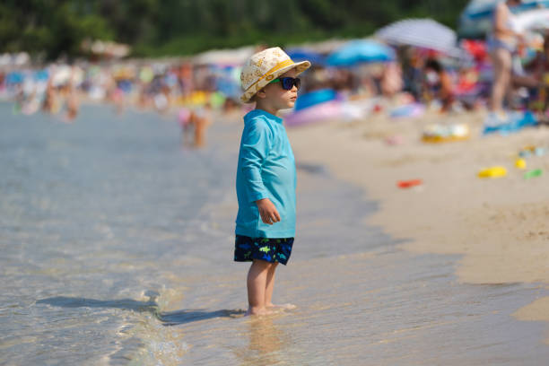 A toddler in a sun hat and blue shirt stands at the water's edge on a crowded sandy beach, suggesting innocence and exploration
