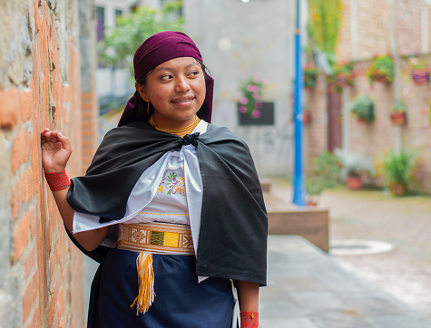 A smiling woman leans against a brick wall, her traditional dress adding a touch of elegance to the quaint, vibrant alley.