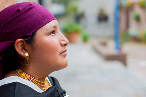 A young indigenous woman from Ecuador wears a vibrant scarf on her head, looks up, lost in thought and adorned in her cultural finest.
