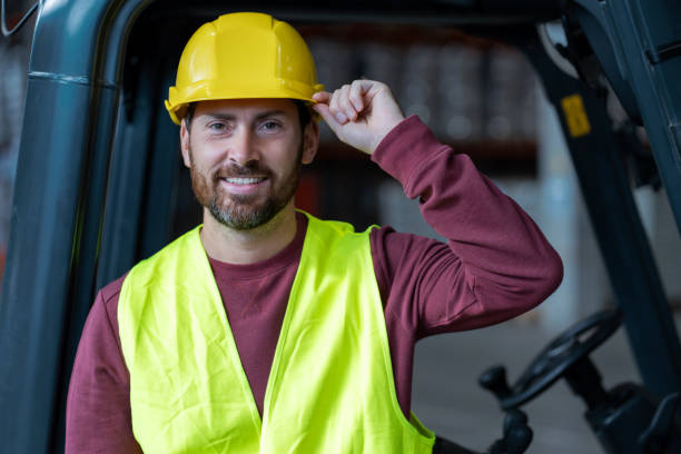 istock Handsome, bearded man, driver, wearing helmet and workwear standing near forklift, looking at camera 2149277417