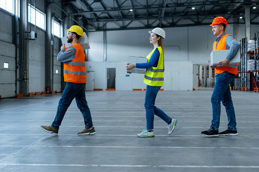 Group of workers, professional loaders, holding boxes, carrying them to warehouse. Young men and woman wearing safety helmet and workwear. Concept of logistics, delivery, distribution