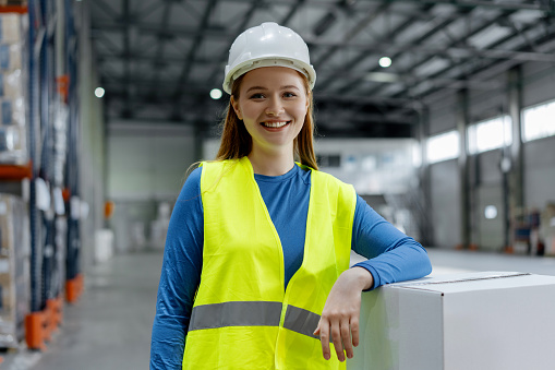 Smiling beautiful young woman engineer wearing hard hat and work wear standing near boxes in warehouse, looking at camera. Concept of logistics, delivery