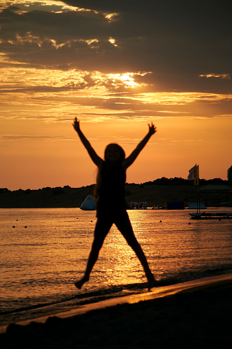 Black silhouette of a female kid jumping in the air in the shape of an X. In the background is a shiny evening sky with beautiful yellow and orange colors