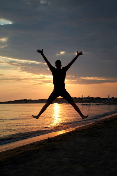 Black silhouette of a young man jumping in the air in the shape of an X. In the background is a shiny evening sky with beautiful yellow and orange colors