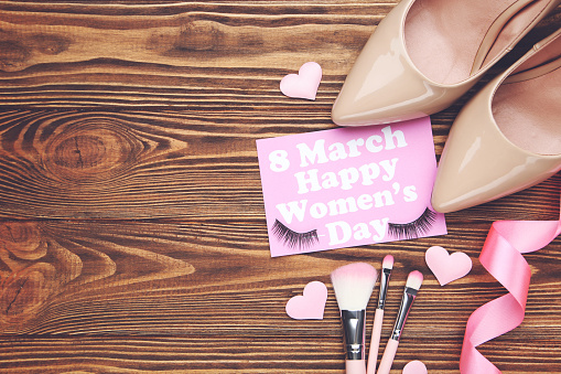 Card with text 8 March Happy Women's Day, pink ribbon and hearts, set makeup brushes and eyelashes, pair beige high-heeled shoes on brown wooden background