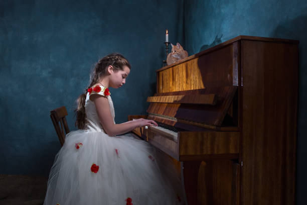 A girl plays the piano. Baby girl in a beautiful dress. Red cat is sitting on the piano. The candle is burning.