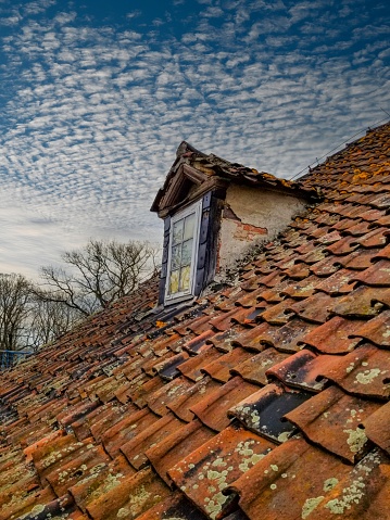 This captivating image showcases a neglected roof window of an old house against the blue sky. The wooden window adds character to the weathered roof with red tiles, creating a nostalgic scene.