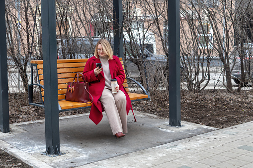 Smiling plus size female blogger in red coat with coffee and phone on swing.