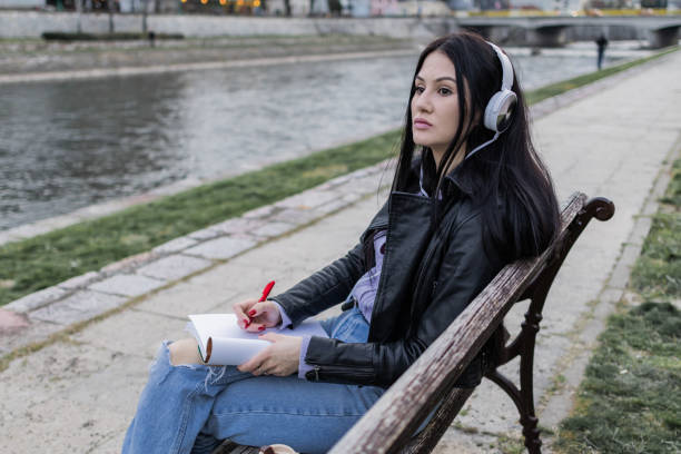 istock Young beautiful female university student with backpack is enjoying near the river. She is wearing headphones and leather jacket while waiting for her college class to start. 2149288737