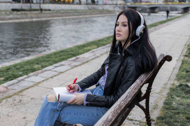 istock Young beautiful female university student with backpack is enjoying near the river. She is wearing headphones and leather jacket while waiting for her college class to start. 2149289196