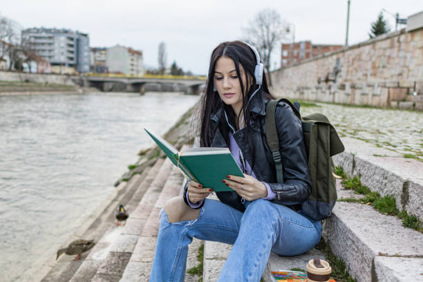 istock Young beautiful female university student with backpack is enjoying near the river. She is wearing headphones and leather jacket while waiting for her college class to start. 2149289258