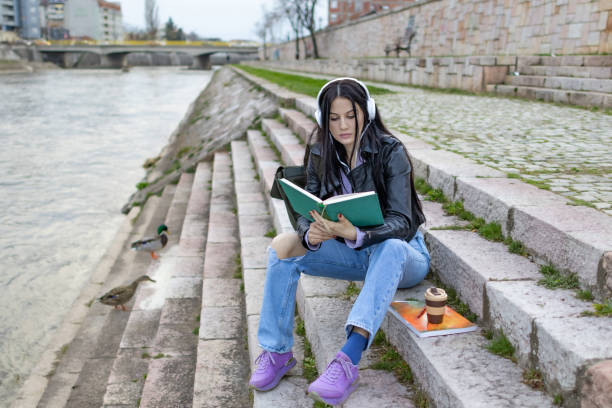 istock Young beautiful female university student with backpack is enjoying near the river. She is wearing headphones and leather jacket while waiting for her college class to start. 2149289588