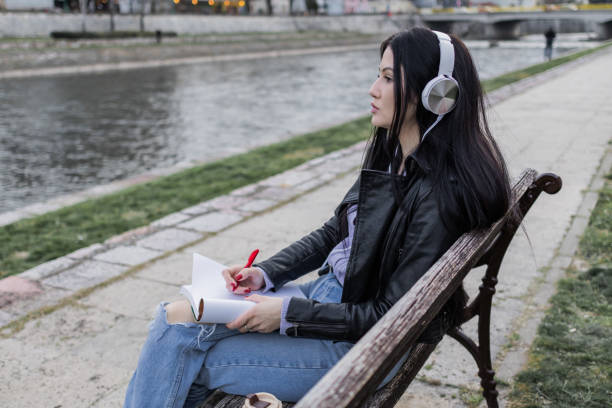 istock Young beautiful female university student with backpack is enjoying near the river. She is wearing headphones and leather jacket while waiting for her college class to start. 2149289632