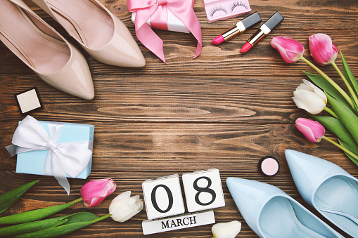 Flowers of tulips, blue and beige high-heeled shoes, gift boxes and cube calendar, lipstick and eyelashes on brown wooden background