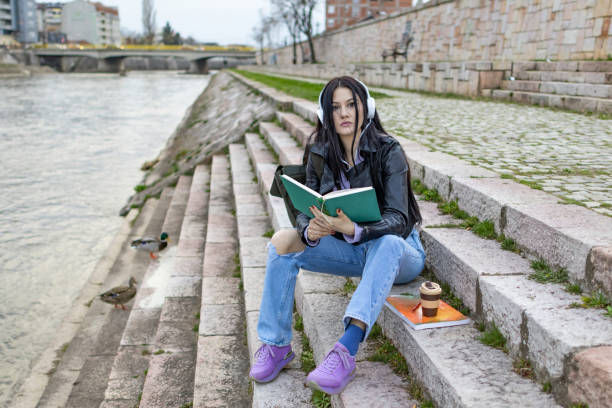 istock Young beautiful female university student with backpack is enjoying near the river. She is wearing headphones and leather jacket while waiting for her college class to start. 2149291165