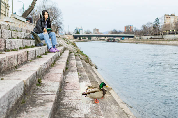 Young beautiful female university student with backpack is enjoying near the river. She is wearing headphones and leather jacket while waiting for her college class to start.