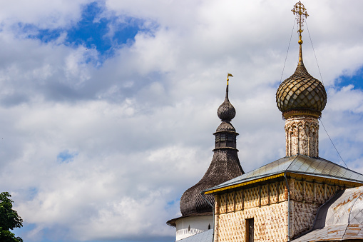 Magnificent scaly dome of an ancient chapel on a round drum next to the hipped wooden roof of a watchtower with a dome and weather vane, details and elements of Orthodox architecture, signs and symbols, sky and clouds, architectural background, history and religion, summer.