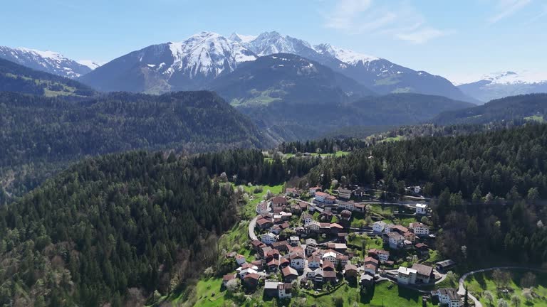 Trin Town in Switzerland surrounded by forest trees in spring. Snowy Alps mountains in background. Aerial forward panorama view.