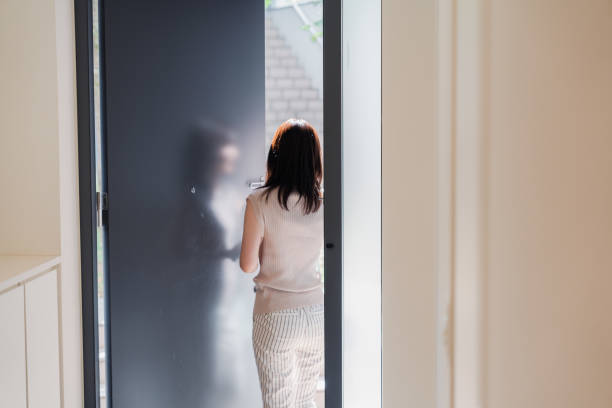 A student seen from behind in patterned uniform, striped sweater, and shoulder bag enters her home by opening a wooden door after returning from a day at school.
