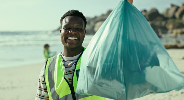 Portrait, beach and black man with a trash bag, environment and climate change with plastic collection and ecology. Face, teamwork or seaside with guy or volunteer with sustainability or eco friendly