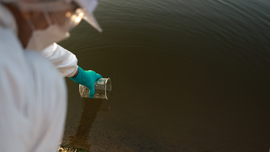 Biologists check the water sample for infections. Gloved hand with Beaker and a test tube on a background of lake, river, sea. Pollution of ecology, environment