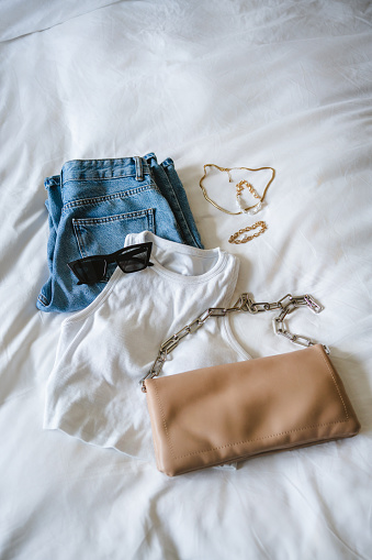 Flat lay with woman's clothes, shoes and accessories: white top, blue jeans, beige leather bag, black sunglasses and gold jewellery. Laid-back yet trendy outfit. Getting dressed and planning outfit concept.