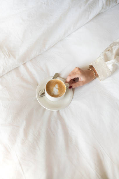 A woman's hand holding a cappuccino on white bed linens