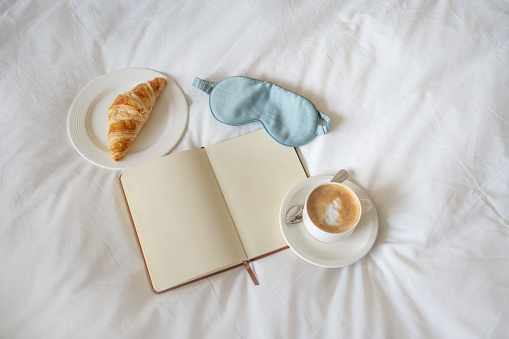 Open journal beside a fresh croissant and cappuccino, with a sleep mask on white bed sheets. Morning journaling concept.