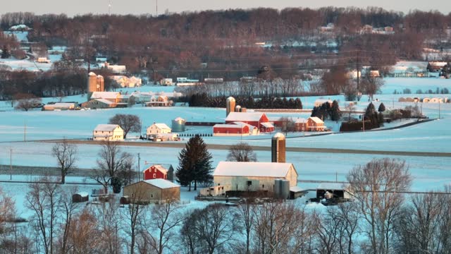 Rural Pennsylvania farmland during winter snow. Aerial parallax shot of farms with barns, houses, and silos during golden hour sunrise on cold snowy day.