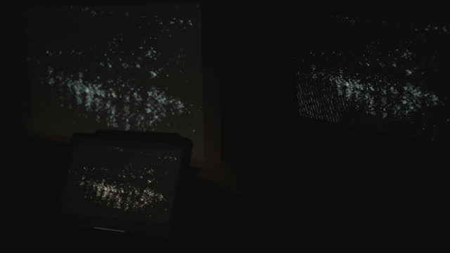 Tight shot of three digital computer devices flashing a strobing static noise signal in a dark room.