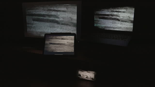 Four digital computer device screens displaying a choppy static noise pattern in a dark room.